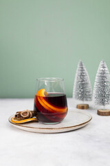 Mulled wine with orange in a glass on a plate on grey and green background.