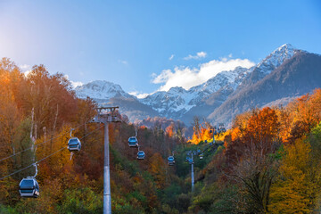 Autumn landscape with mountain range green, yellow red foliage of forest trees illuminated by the sun. High mountains with snow capped peaks with a cable car, and passenger cabins rising up.