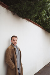 Young fashionable man in long brown coat and scarf leaning on white wall on the street, hands in pockets, looking away, mid-thigh portrait crop, plenty of copy space left