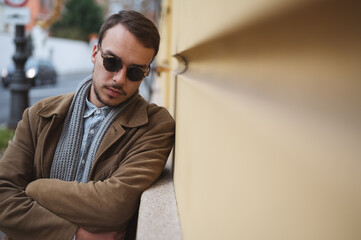 Young street fashion model with sunglasses and brown coat leaning on building wall, looking down