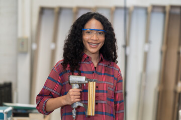 Smiling young woman carpenter holding pneumatic nail gun at wood workshop. Portrait of joiner...