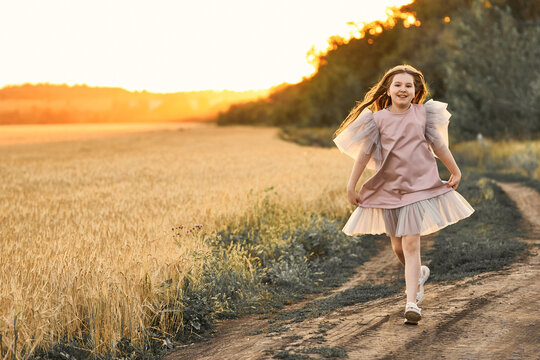 Cute emotional smiling girl with long hair cheerfully running in the field in sunset at a summer evening. Healthy summer activity for children. Having fun.