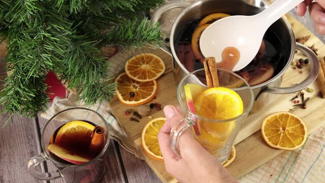Christmas punch in pot, pouring flavored mulled wine in glasses, alcohol punch scented with spice and cinnamon, stewed fruit pot placed on wooden table, slow motion 4k video footage