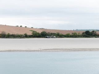 Sandy seashore on a cloudy day. Calm water surface. The remains of an old ship on the shore. A plowed field. Minimalistic landscape.