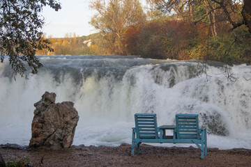 Two jointed blue wooden chairs under tree shade at the river shore affront of small water jumps,...