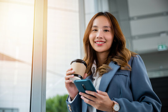 Young Asian businesswoman wearing blue shirt holding coffee cup and her smartphone. business woman smiling standing holding mobile phone near window and elevator blur background in the office.