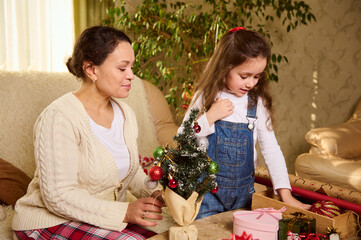 Happy family making New Year's preparations. Loving mother and her adorable little girl, pretty daughter, packing presents and decorating Christmas tree, in anticipation for the upcoming winter events