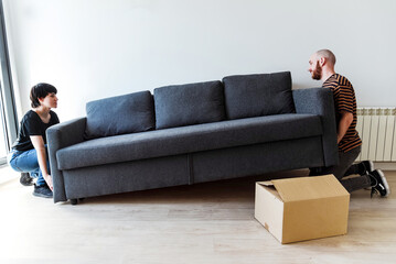 Una pareja joven levantando un sofá para moverlo. Concepto de amueblar una nueva casa. Hipoteca. Independizarse. - obrazy, fototapety, plakaty