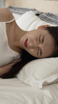 Vertical Screen: asian girl having severe cramp in lower abdomen is holding her belly and lying on bed with a groan of pain in the bedroom at home during daytime.