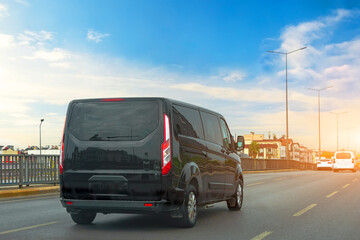 Black modern passenger charter van with a small delivery moves fast on the highway to the urban suburbs. Business distribution and logistics express service. Mini bus rides along the highway. - obrazy, fototapety, plakaty