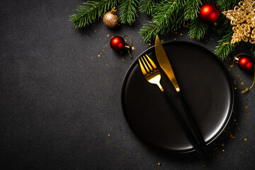 Christmas table with black plate, golden cutlery and holiday decorations. Top view on black.