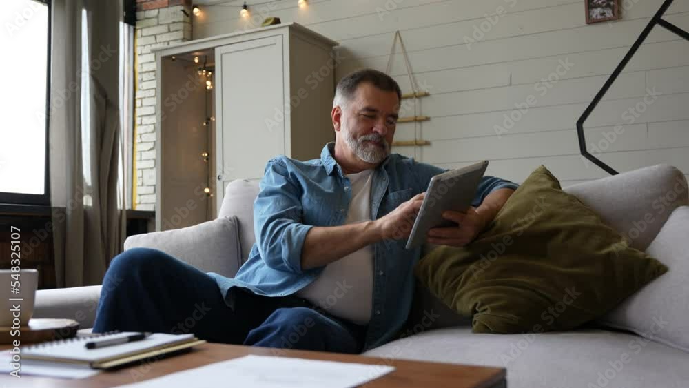 Wall mural Senior internet user. Positive mature grey-haired man browsing online on digital tablet, networking or playing video games, sitting at home