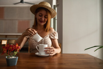 Middle aged woman in straw hat pours milk into cup with coffee or tea at table, resting in summer cafe with tropical flowers, sitting indoors. Travel tourism and cooking concept. Copy text space - obrazy, fototapety, plakaty