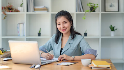 Asian businesswoman sitting happily with her laptop and takes notes intensely and smiles happily at her assignment.