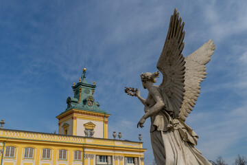 the statue of an angel holds a wreath in his hand directed towards the Wilanowski Palace in Warsaw, the statue is against the background of the blue sky, a view from below
