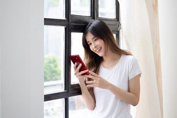 Portrait of attractive asian woman in casual clothes is using a smart phone and smiling while standing near the window, holding mobile phone.