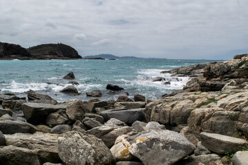 Grandes rochas da praia das Conchas, próxima a cidade de Cabo Frio, com mar azul em volta, céu com nuvens, rochas e montanhas ao fundo.