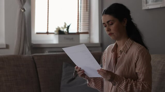 Sitting independent young Caucasian woman reads lease or purchase agreement and roll key on finger. Loan and rental apartment. Concept of leasing and mortgage.