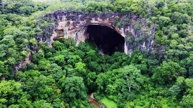 Big open cave in lush green tropical forest with small crystal clear stream running into the cave camera approaching cave obove trees Aerial Video