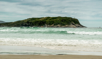Orla do Chateau Recanto das Dunas na Praia das Dunas, com ondas fortes, pequenos barcos e farol encima de rochedo.