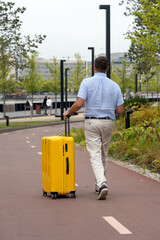 Fototapeta na wymiar Back view of millennial man in casual wear carrying his yellow luggage while walking through city park