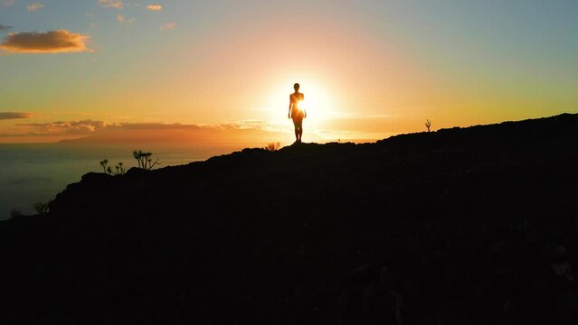 Woman silhouette on mountain top at sunset light aerial drone flight. Sun rays otline a person. Hiker enjoying sunrise. Sunbeams rise from ocean.