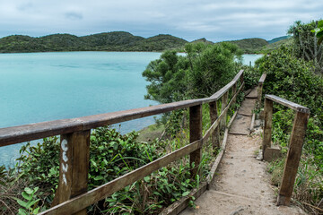 Ponte de madeira perto do Forte São Mateus em Cabo Frio, com mar azul e muitas montanhas ao fundo.