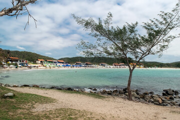 Vista da linda praia das Conchas, próxima a cidade de Cabo Frio, árvores e quiosques a beira-mar, céu azul, mar com águas limpas e em tom de azul, com montanhas ao fundo.