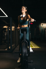 Fototapeta na wymiar An Asian woman in a gym exercising on a bicycle. Women making an effort while exercising on an air bike. The girl is wearing headphones.