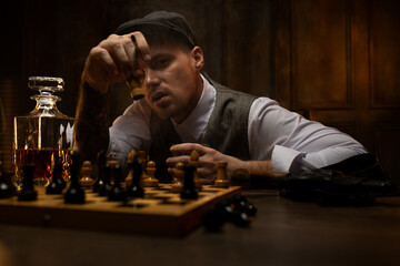 Young gangster drinking whiskey and playing chess at table in dark pub