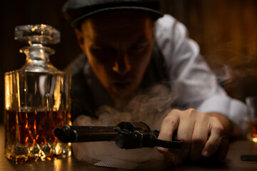 Revolver in hand of gangster leaning on table with cards and decanter of whiskey