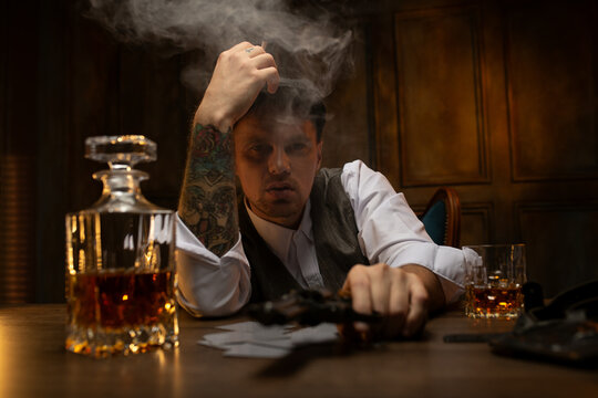 Young gangster smoking and holding revolver at table with cards and decanter of whiskey