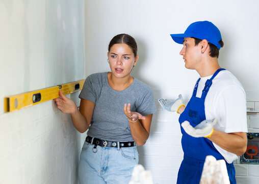 Girl dissatisfied with quality of renovation works carried out in her apartment, checking flatness of wall surface with spirit level and talking to confused young builder..