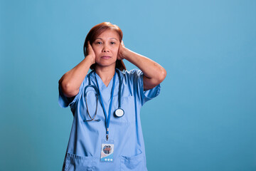 Asian quiet nurse with stethoscope covering ears having serious expression during checkup visit...
