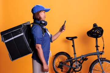 Slide view of takeaway delivery worker standing beside bike while checking directions and order details on smartphone. Restaurant worker wearing delivery uniform while carrying takeout food backpack