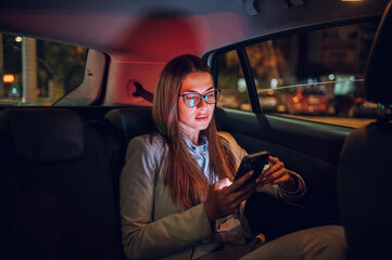 Business woman using smartphone while sitting in a backseat of a car at night