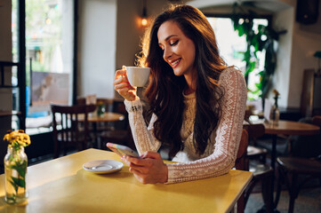 Portrait of beautiful young woman using smartphone and drinking coffee in a cafe
