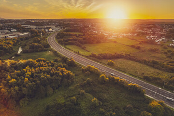 Road in town by a green park and residential area with homes at warm sunset. Sunset time. Stunning nature scenery in the background. Galway city, Ireland. Aerial view. City transportation system. - obrazy, fototapety, plakaty