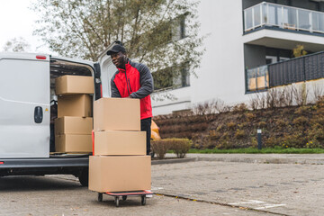 Full-length outdoor shot of African-American delivery man using hand truck to deliver cardboard boxes. White delivery van with opened trunk full of packages. Modern building block in the background