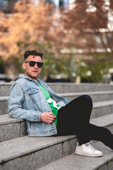 Young blonde model guy with sunglasses, green t-shirt, denim jacket and black jeans sitting on stone stairs in a park in the city in the  afternoon
