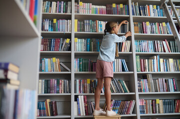 Girl standing on the chair and taking a book from the shelf