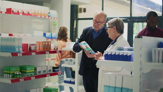 Senior man asking specialist about healthcare treatment, reading pharmaceutical leaflet to buy medicaments. Pharmacist giving box of cardiology pills to client, helping with disease. Tripod shot.