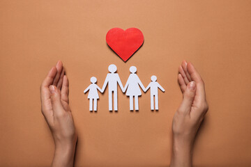 Woman protecting paper family figures and red heart on brown background, top view. Insurance concept
