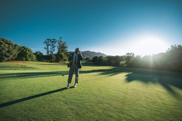 Fototapeta premium A wide-angle view of a dashing bald African American man in an elegant tailored outfit adjusting his sunglasses while staying on the lawn of a golf field with a club in his hand on a sunset
