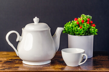 White plain ceramic teapot and cup with red holly berries potted plant on a wooden table with a black background