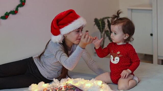 Beautiful caucasian brunette girl in a santa claus hat eats a candy from the advent calendar lying on the bed resting her head on her hand, and the younger sister asks pointing her finger and the olde