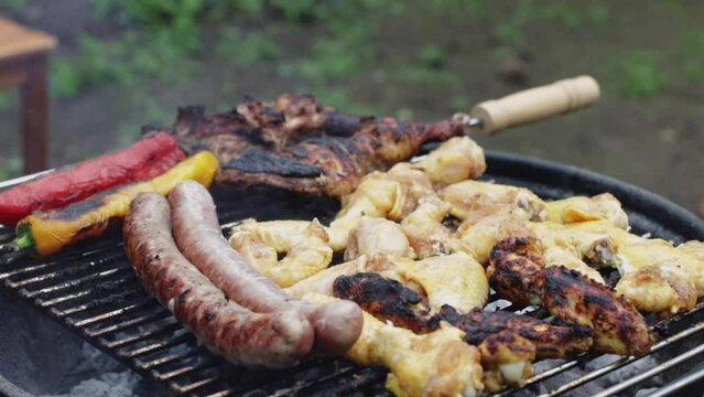 Pouring water over hot juicy fried chicken wings, sausages and steak on a grill with pepper fire and smoke, standing in the backyard of his house, close-up side view in slow motion with zoom out.