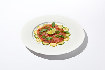 Beef carpaccio with zucchini and cucumber on white table with sunlight shadows. Appetizer of meat - beef carpaccio on summer dining. Sliced raw meat in italian style. Aesthetic menu.