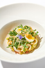 Healthy salad with squid and avocado on white plate with harsh shadows. Calamari salad with vegetables and eggs on white background with shadows of leaves. Summer food menu.