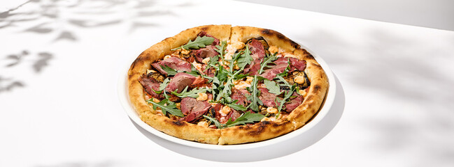 Italian pizza with roast beef, arugula and spicy sauce on light background with shadows. Meat pizza with sliced roast beef on white table. Italian food in trendy menu restaurant